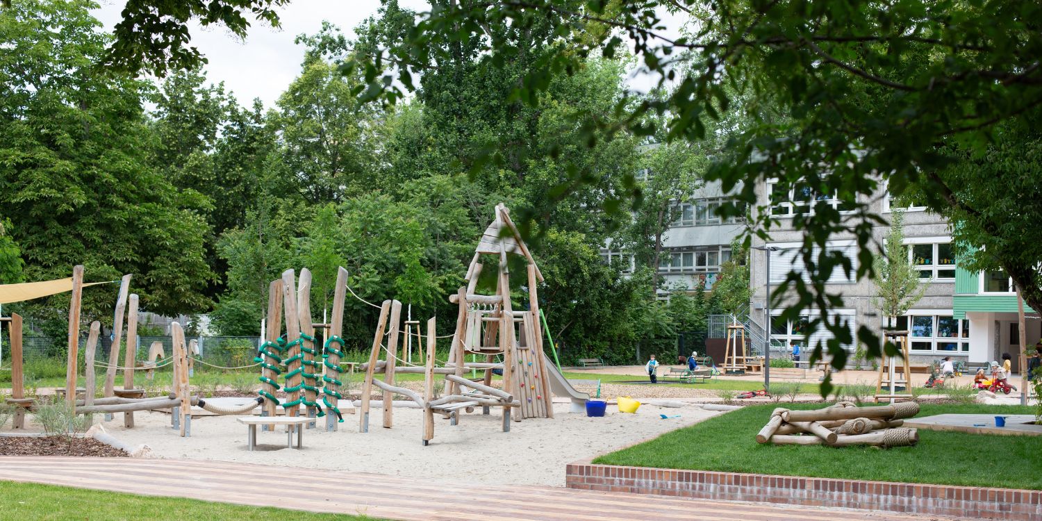 Holzgeräte und Holzklettergerüste stehen unter großen Sonnensegeln im Spielsand. Im Hintergrund steht das Kitagebäude.