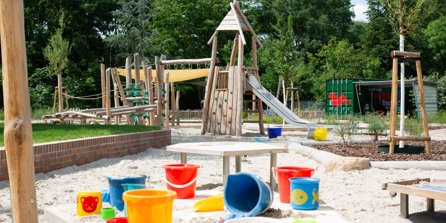 Im Sandkasten der Kita steht ein Klettergerüst aus Holz. Im Vordergrund liegen bunte Buddeleimer.