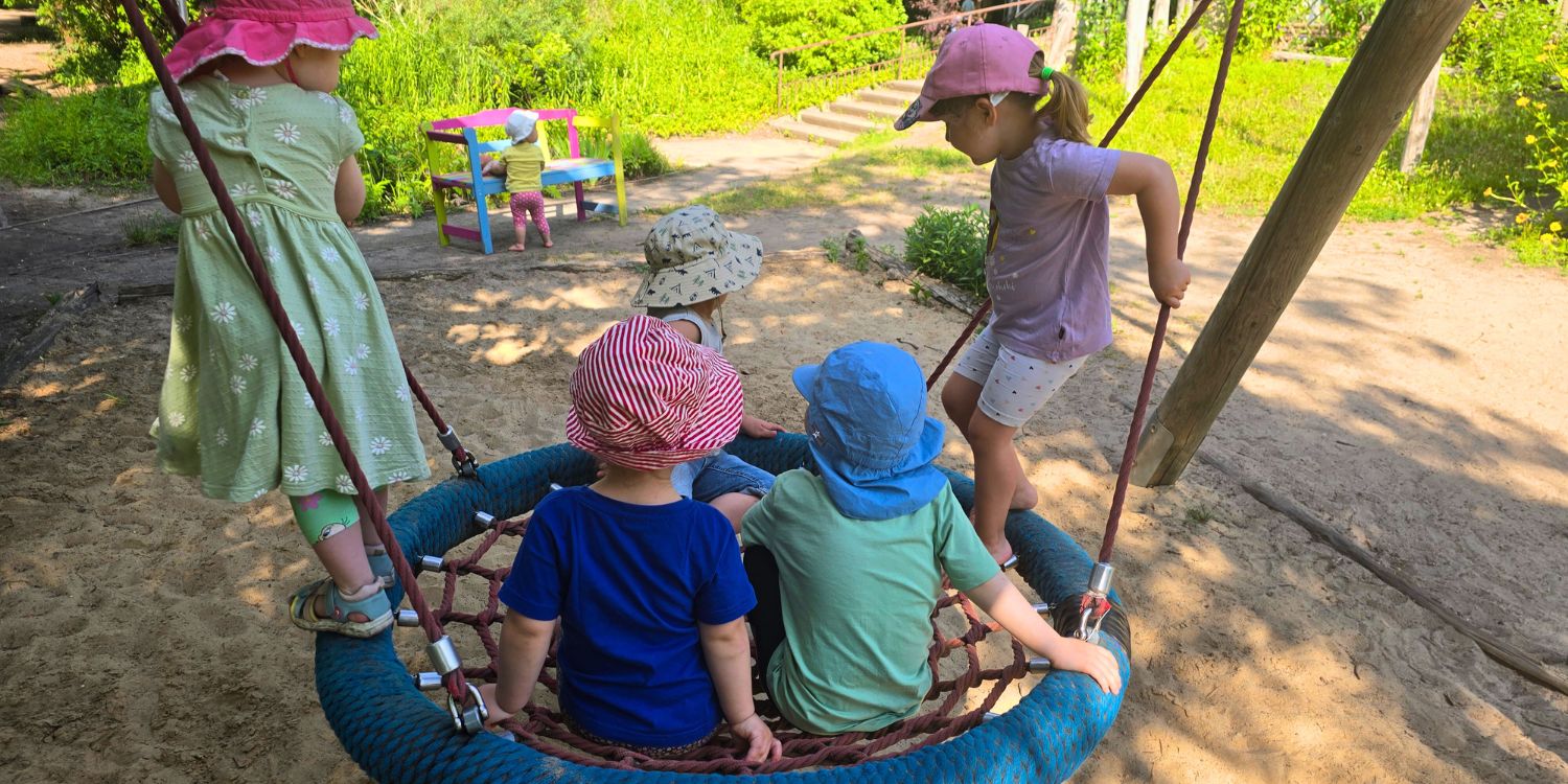 Drei Kinder sitzen in einer Nestschaukel, zwei stehen am Rand. Im Hintergrund steht ein Kind vor einer Bank.