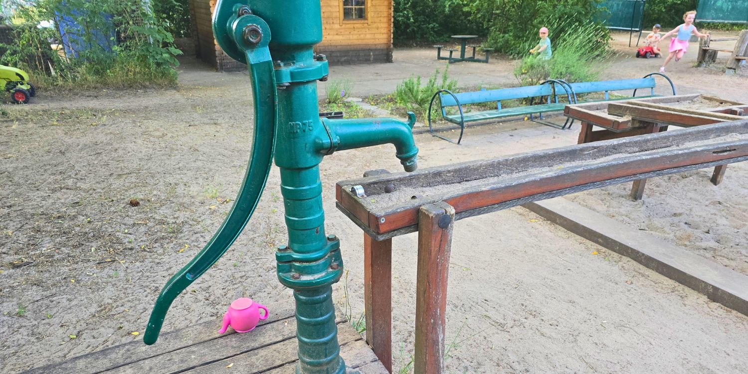 Eine Wasserpumpe führt zu einer Matschecke mit verschiedenen Wasserläufen aus Holz. Im Hintergrund laufen Kinder und fahren mit dem Dreirad.