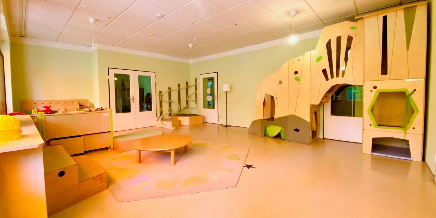 In einem großen Gruppenraum stehen verschiedene Holzspielzeuge und Klettermöglichkeiten. An der Wand hängt eine weitere Klettermöglichkeit in Form eines Tieres.