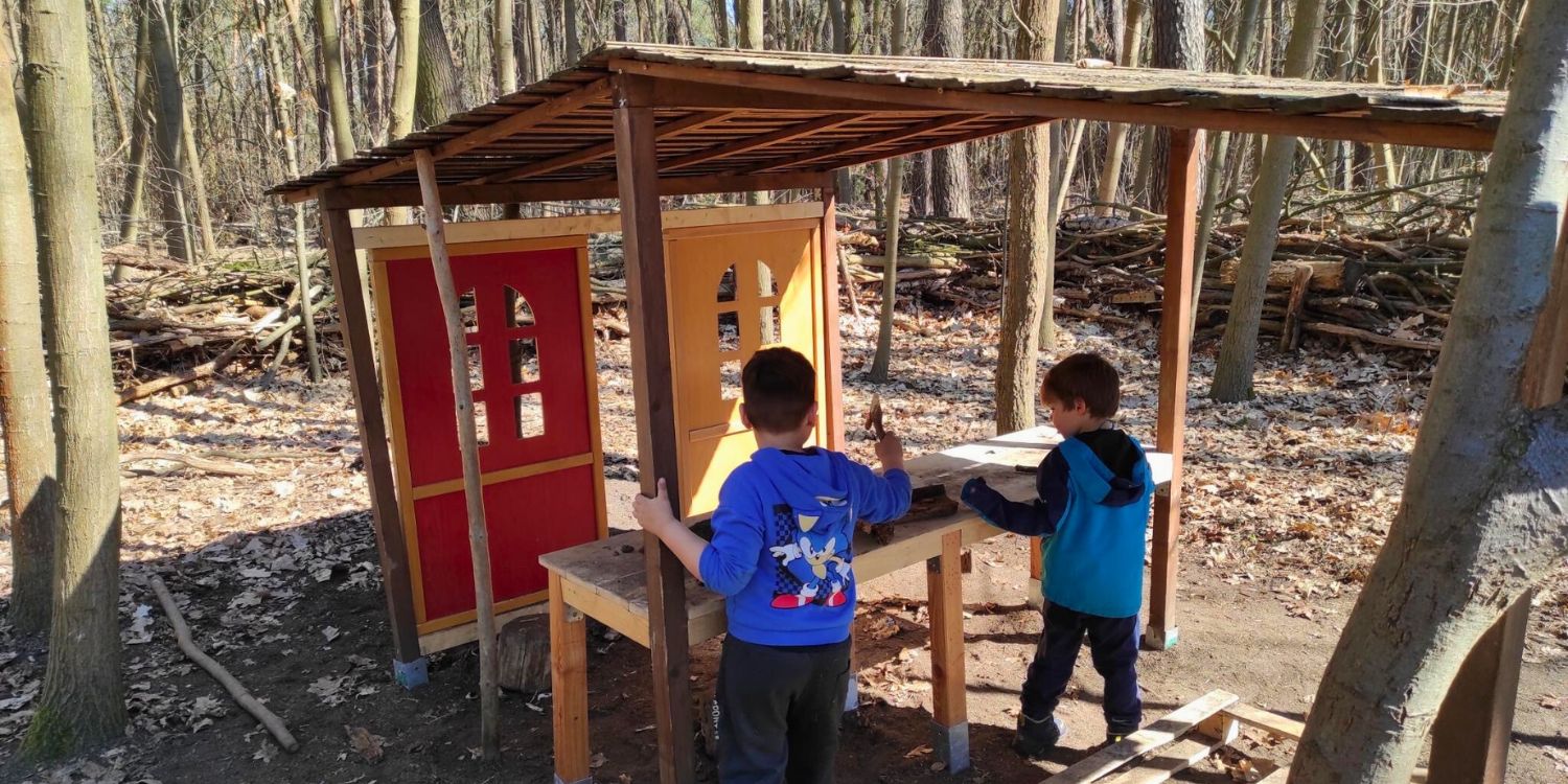 Zwei Jungs stehen in einer aus Holz gebauten kleinen offenen Hütte, die eine Waldküche darstellen soll.