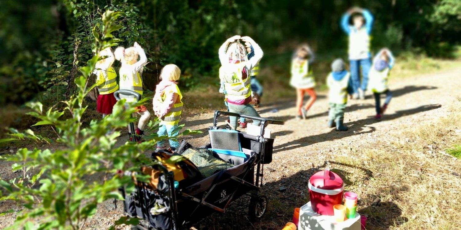 KInder gehen mit ihrer Erzieherin durch den Wald. Im Vordergrund steht ein Bollerwagen mit Verpflegung und Geschirr.