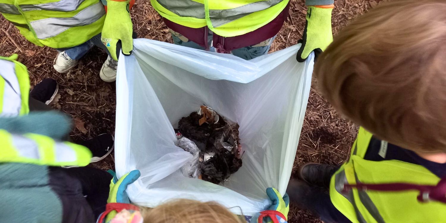 Kinder mit Handschuhen halten einen Müllsack offen, so dass alle sehen können, was sie im Wald gesammelt haben.