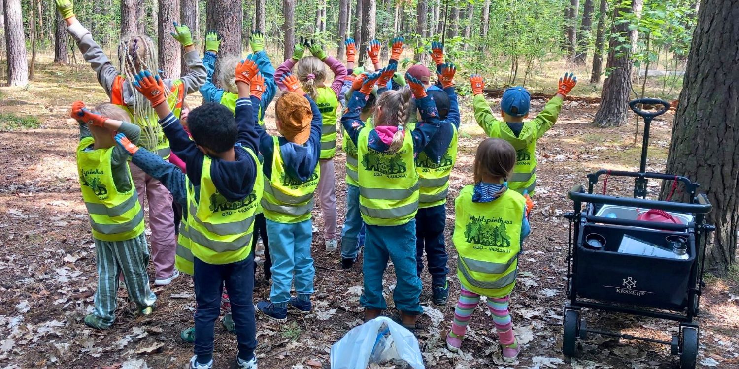 Ein Gruppe von Kindern steht im Wald und hebt die Hände empor. Rechts im Bild steht ein Bollerwagen.