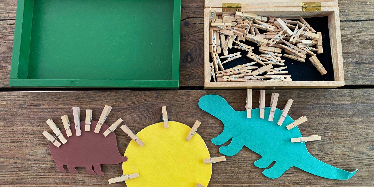 Auf einem Tisch liegen aus Papier ausgeschnitte Figuren - eine Sonne, ein Igel und ein Dino. An ihren Rändern klemmen Wäscheklammern, Darüber liegt eine Kiste mit Wäscheklammern und eine leere Kiste.