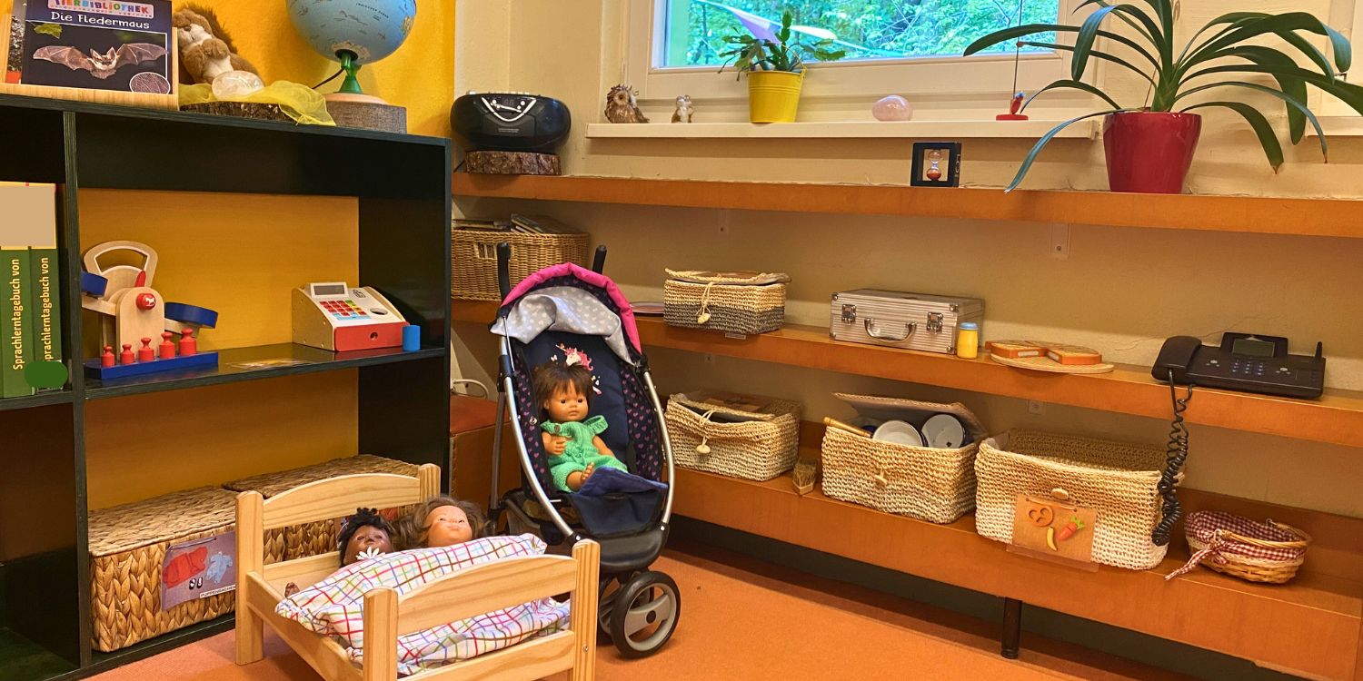 In der Ecke eines Raumes stehen ein Puppenbettchen und ein Puppenwagen. An den Seiten stehen Regale mit Büchern und Bastelmaterial.