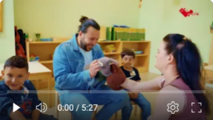 Ein Mann sitzt auf einem Stuhl und hält eine Handpuppe in der Hand. Vor ihm sitzt eine Frau, die ebenfalls eine Handpuppe in der Hand hält. Neben ihnen zwei Kinder, die zusehen