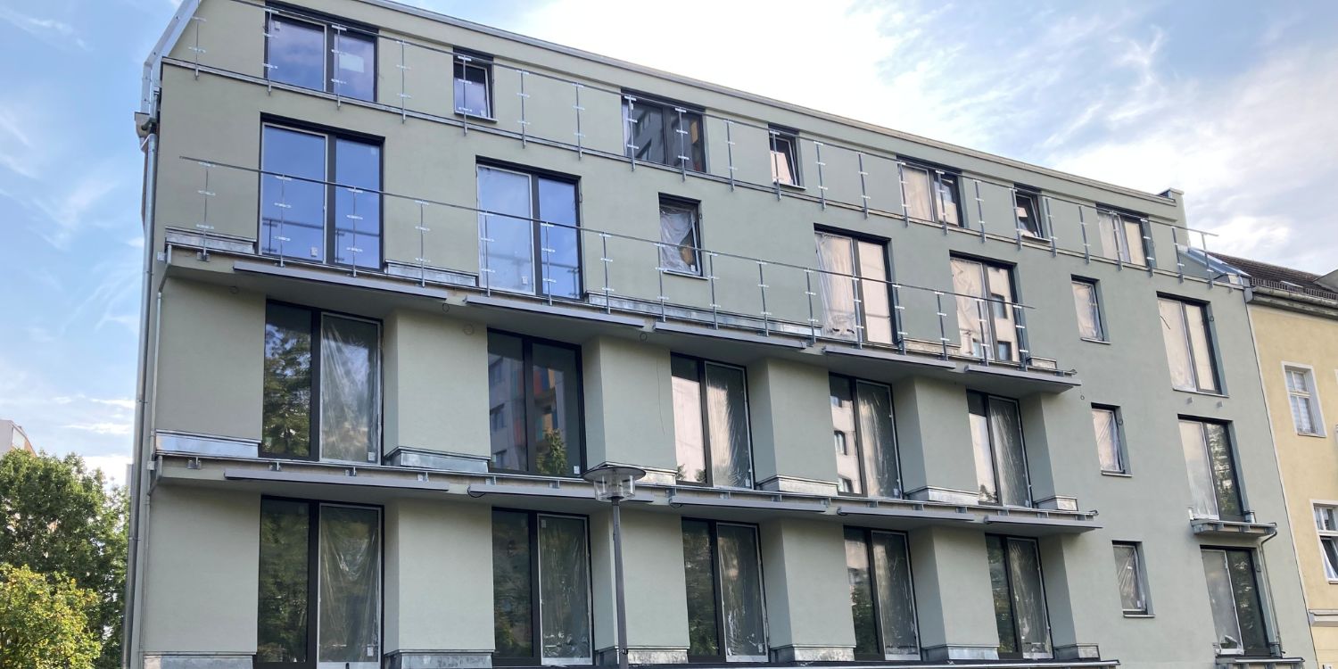 Ein graues Haus mit vier Etagen steht in einer Straße; im Hintergrund sind Bäume zu erkennen.